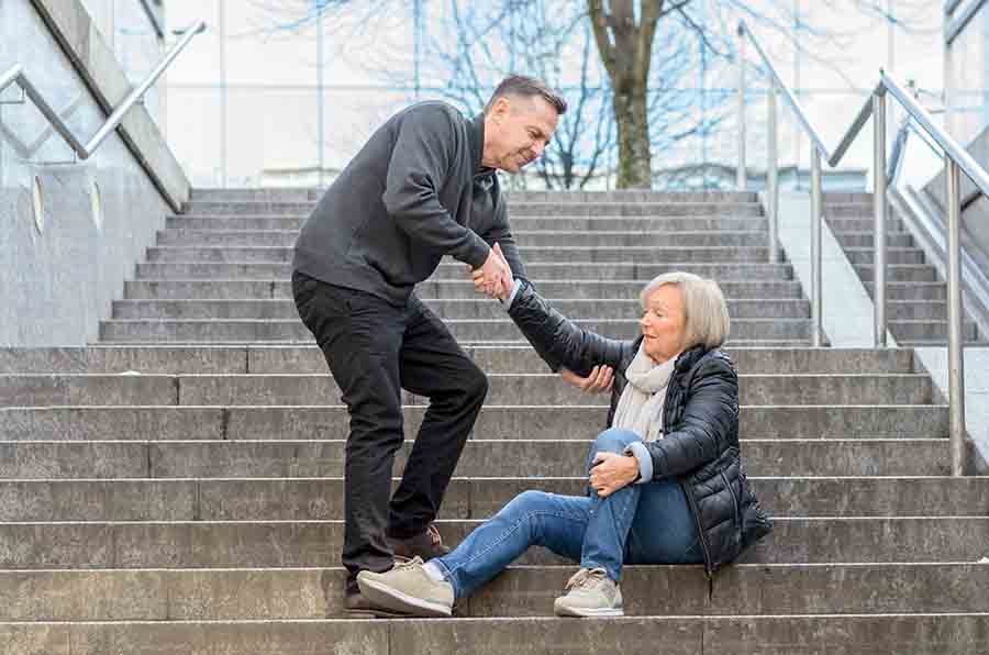 Man bending down to help a woman clutching her knee get up after a fall on stairs outside