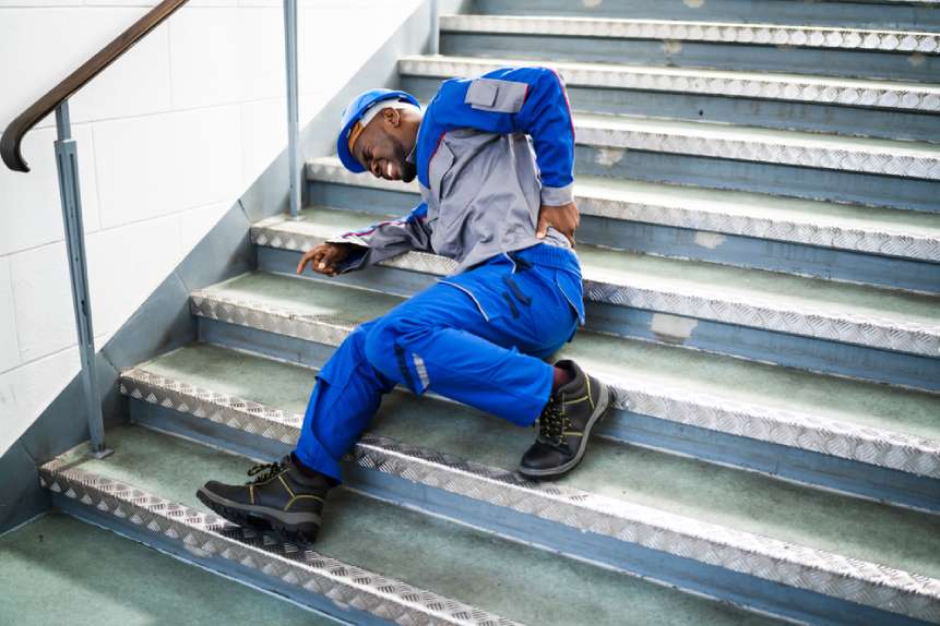 Trabajador tumbado en un escalón agarrándose la parte baja de la espalda y haciendo muecas tras resbalarse y caerse en un accidente laboral.
