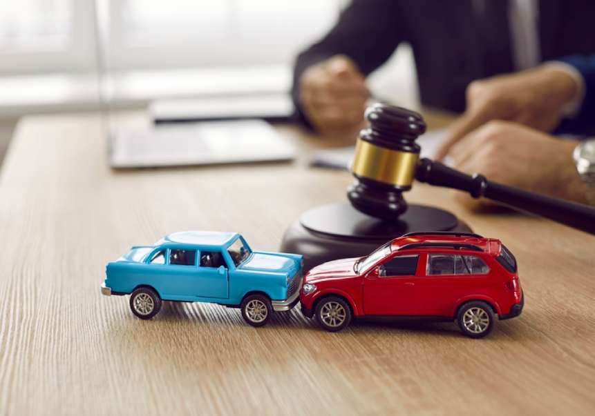 Coche de juguete azul pequeño y coche de juguete rojo pequeño choque frontal sobre mesa con mazo y abogados de fondo.
