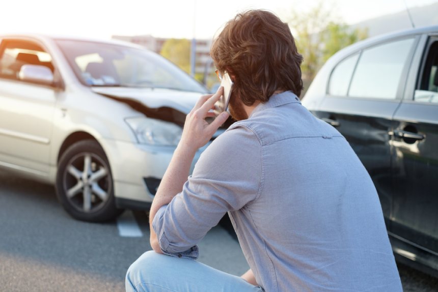 Man in shirt and jeans crouching on ground facing car accident scene using phone