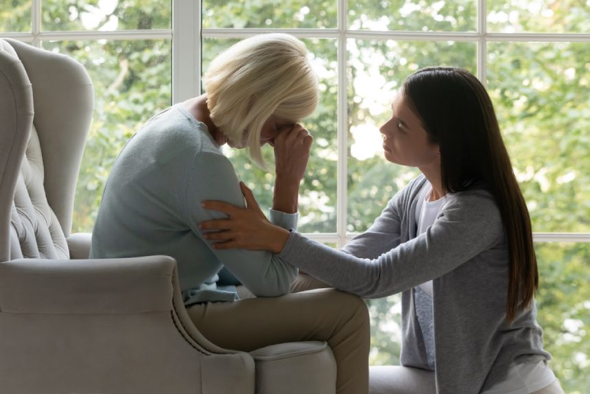 Young woman kneeling before grieving senior woman and comforting her after bereavement