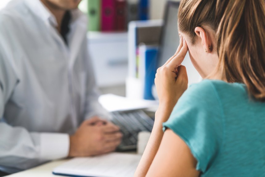 Mujer rubia con dolor de cabeza frente a médico en consulta masajeando sienes