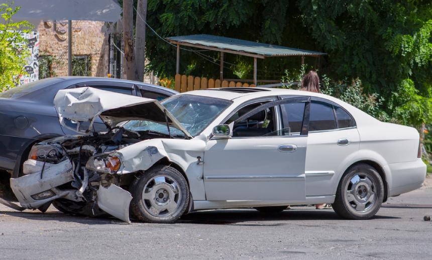 Two smashed cars light gray and black with woman standing behind gray car in background