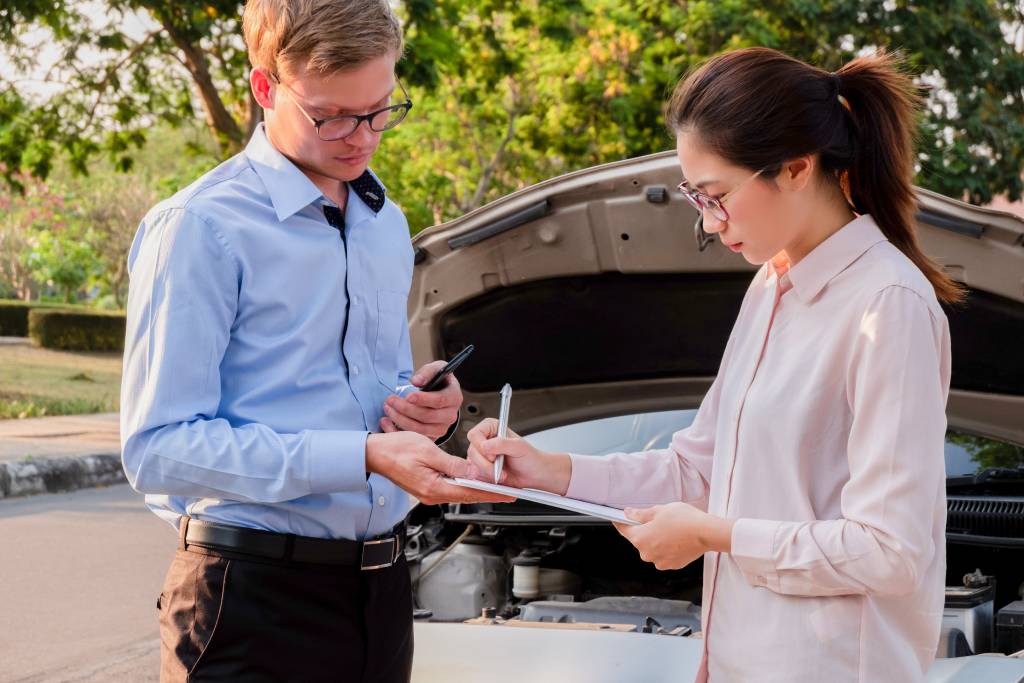 Young insurance adjuster and car accident victim completing paperwork in front of damaged car