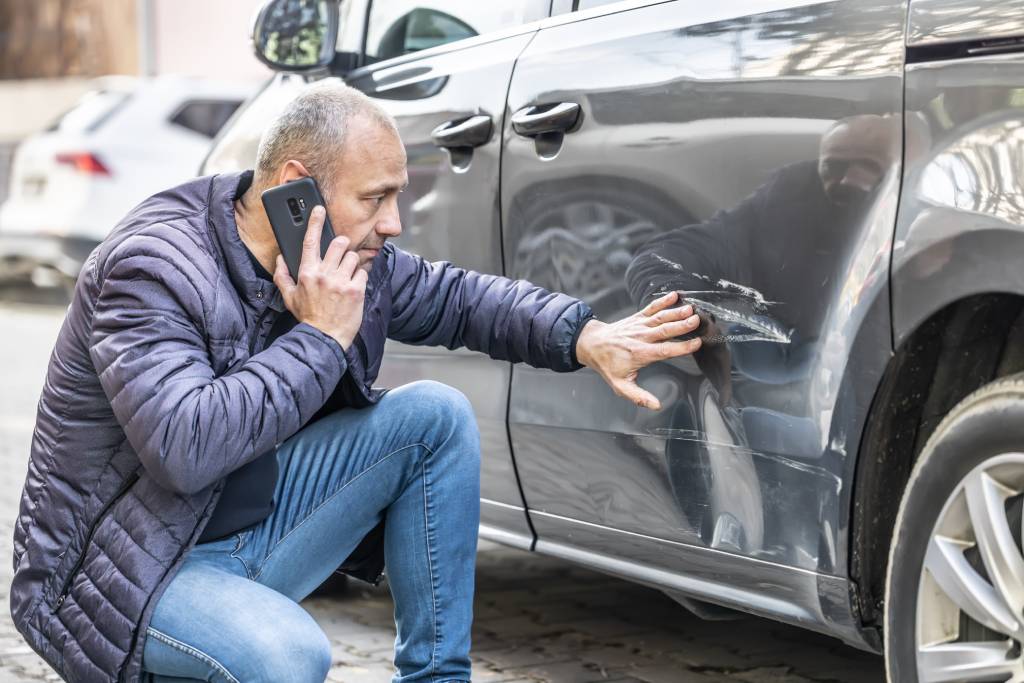 Man on phone crouched beside vehicle inspecting damage to left rear side panel