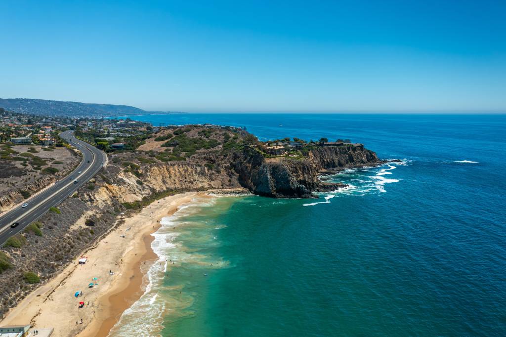 Aerial view of roadway snaking towards beach with ocean and cliffs on the right