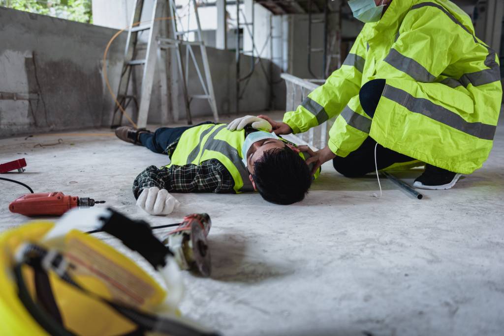 A construction worker in a safety vest lies injured on a concrete floor while another worker attempts to help.