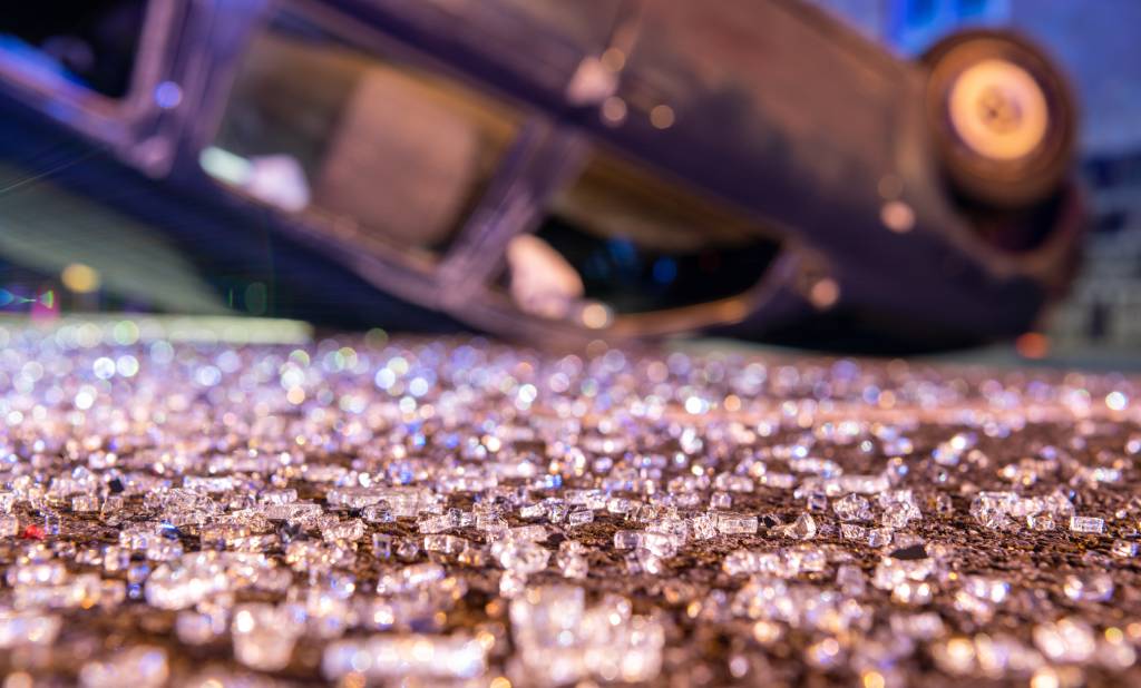 A vehicle rollover accident, the car blurred by depth of field but easily recognized. Hundreds of shards of glass remain on the pavement.