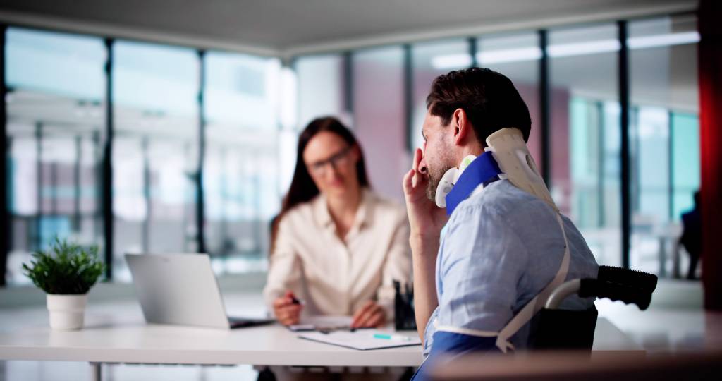 A man in a neck brace in the foreground winces in pain while speaking to an HR manager or lawyer in an office.