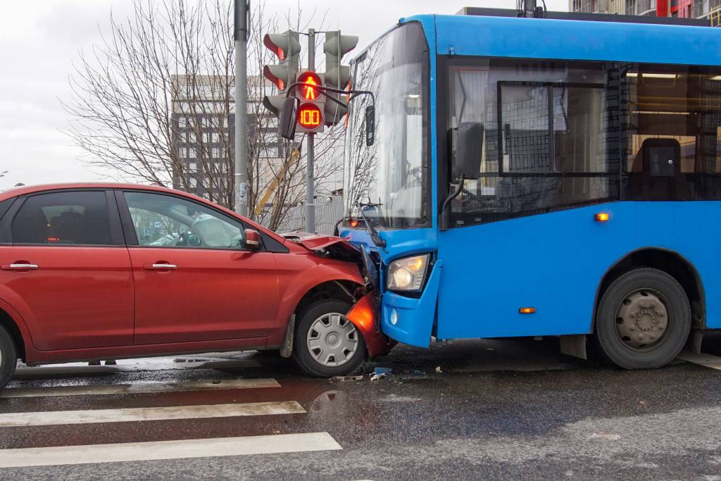 Un turismo rojo choca frontalmente contra un autobús de pasajeros azul, con un semáforo rojo sobre los dos vehículos.