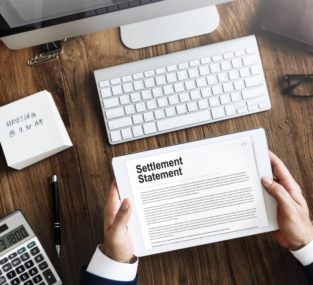 A lawyer reads a settlement statement on a tablet while sitting at a desk that also features a desktop computer, monitor, notepad, etc.