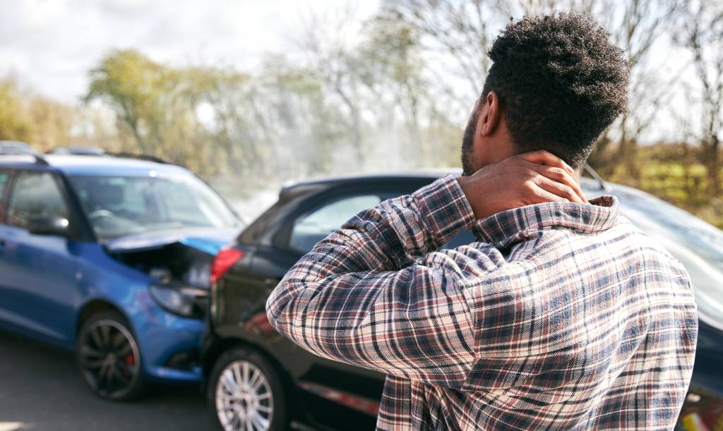 A young man in pain from whiplash rubs his neck while standing by his damaged car after a traffic accident.