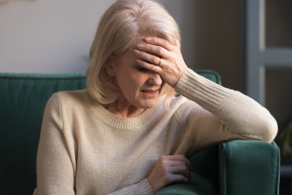 Woman with light blonde hair grimacing and clutching her forehead in pain sitting on sofa